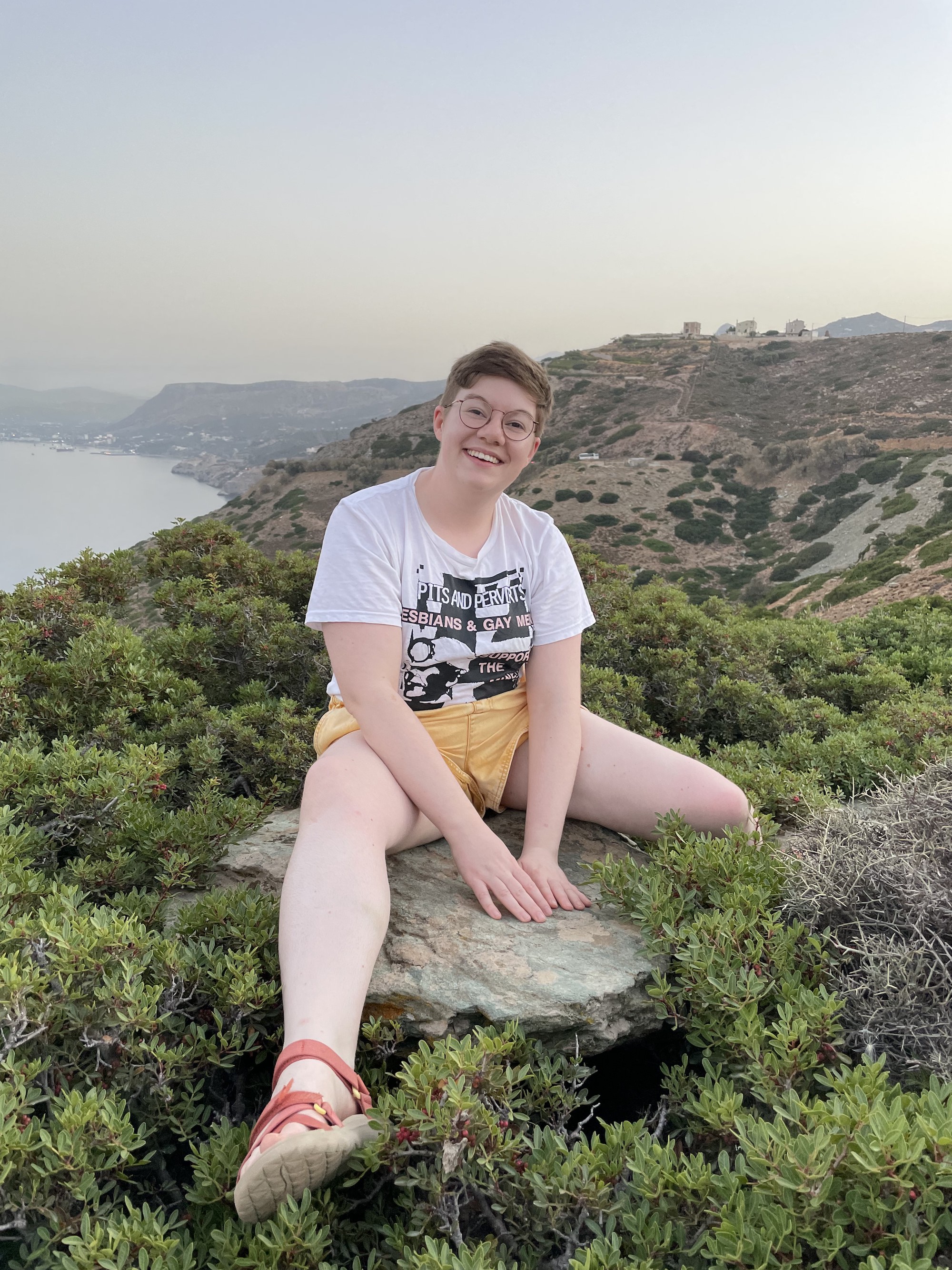 A white non-binary butch with short, brown hair and glasses. They're wearing yellow shorts and an LGSM t-shirt. They are sitting on a rock on the top of a hill by the ocean.
