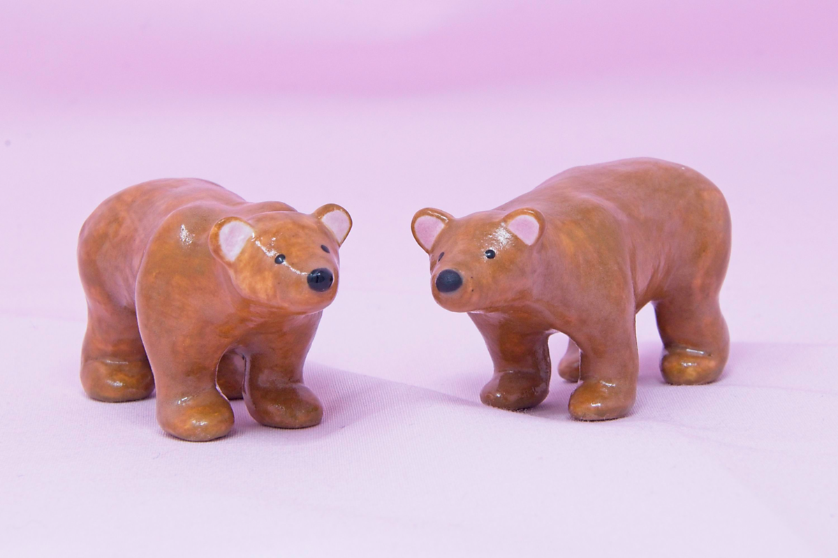 A pair of handmade miniature ceramic brown bears against a pink background. They're posed to face each other.