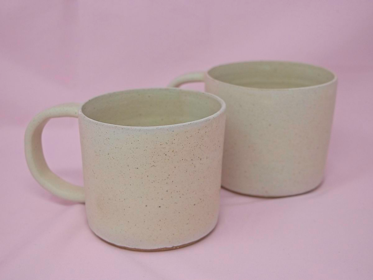A pair of handmade cream speckled mugs against a pink background.